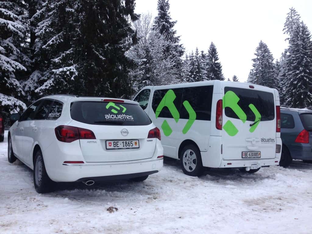 Deux voitures avec le logo d'Alouettes Cycles, parker sur de la neige avec en fond des sapins enneigés.